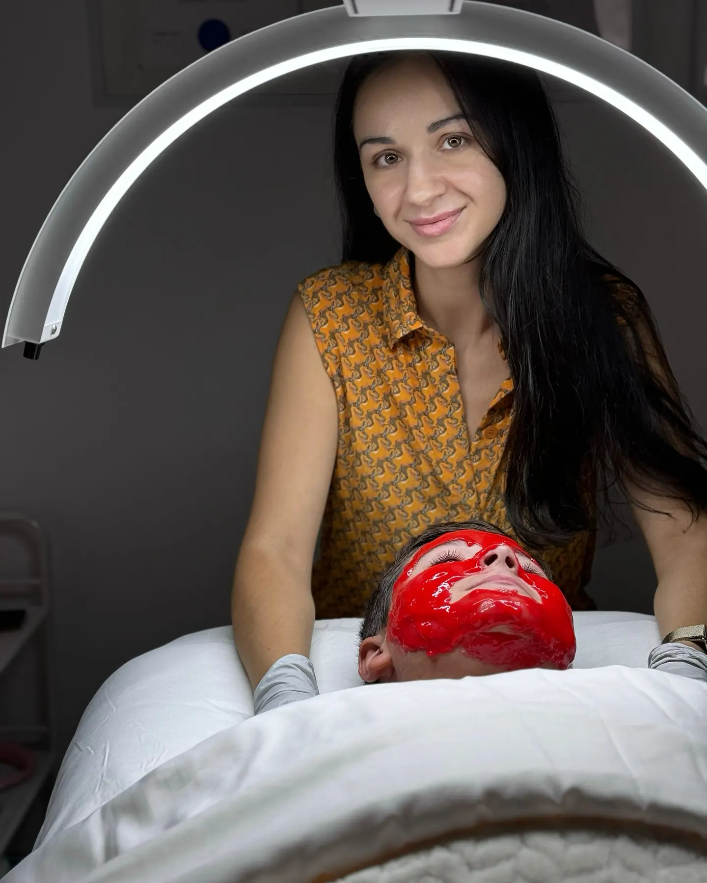 Woman in orange shirt applies red mask to person's face.