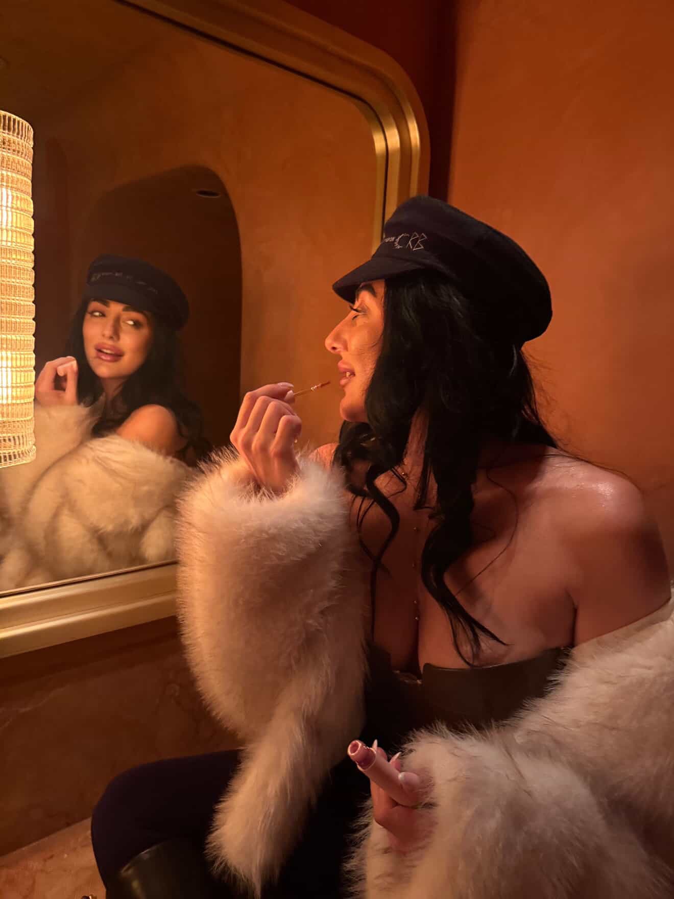 Woman in fur coat applies lipstick in front of mirror.