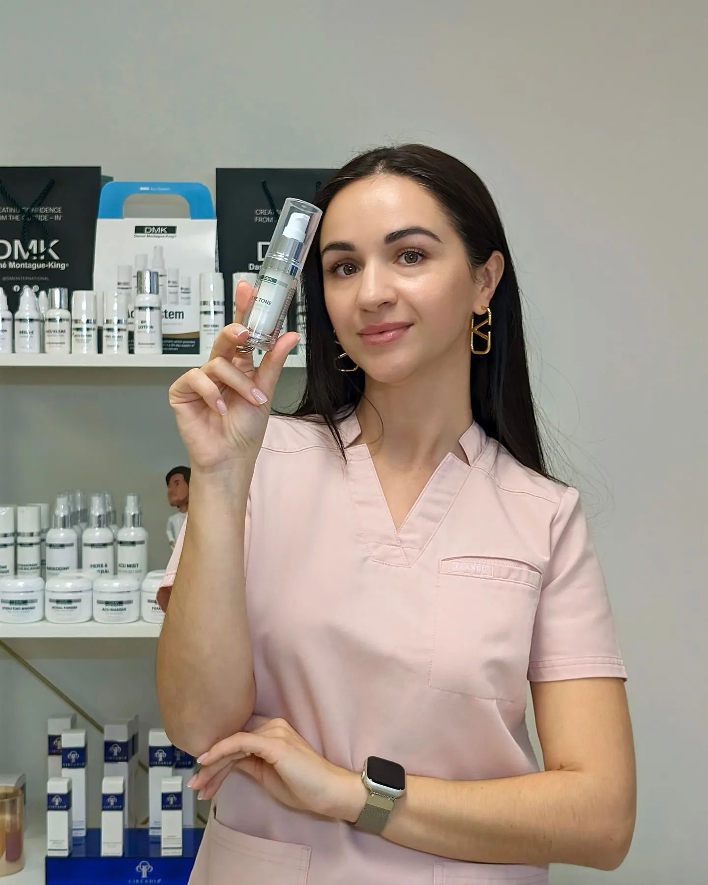Woman in pink scrub holds DMK skincare product in front of shelves.